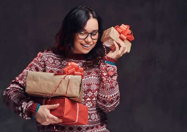 Portrait of a happy brunette girl wearing eyeglasses and warm sweater holding a gifts boxes, isolated on a dark textured background. — Stock Photo, Image