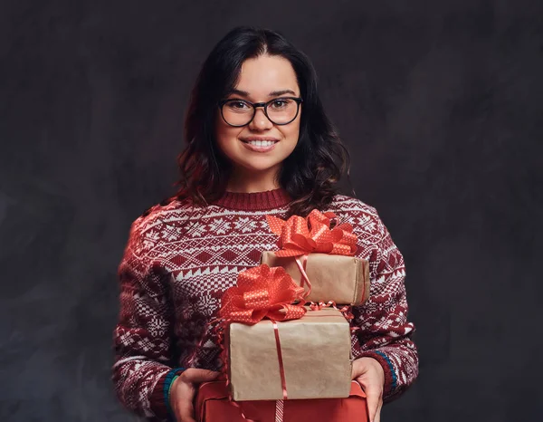 Portret van een gelukkig brunette meisje dragen van bril en warme trui houden een geschenken-dozen, geïsoleerd op een donkere gestructureerde achtergrond. — Stockfoto
