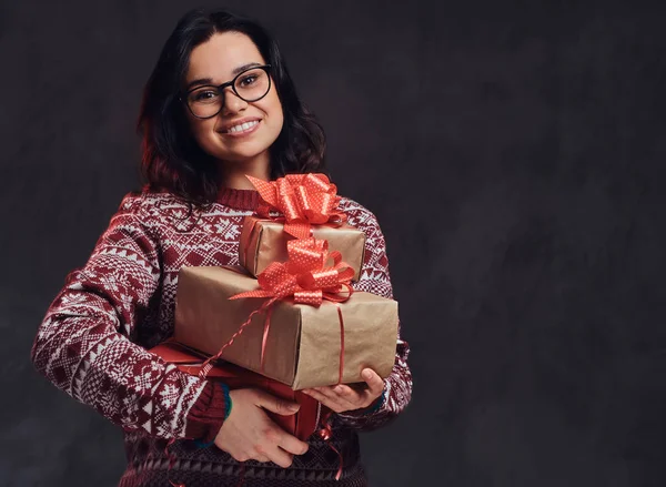 Portret van een gelukkig brunette meisje dragen van bril en warme trui houden een geschenken-dozen, geïsoleerd op een donkere gestructureerde achtergrond. — Stockfoto