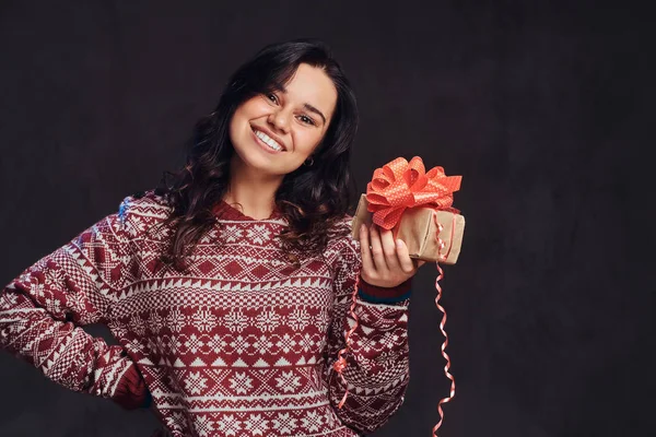 Retrato de uma menina morena feliz vestindo uma camisola quente segurando uma caixa de presente, isolada em um fundo escuro texturizado . — Fotografia de Stock