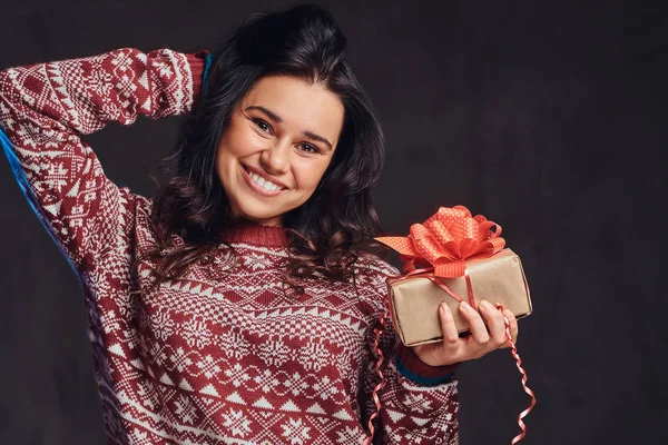 Portret van een gelukkig brunette meisje draagt een warme trui die houden van een doos van de gift, geïsoleerd op een donkere gestructureerde achtergrond. — Stockfoto