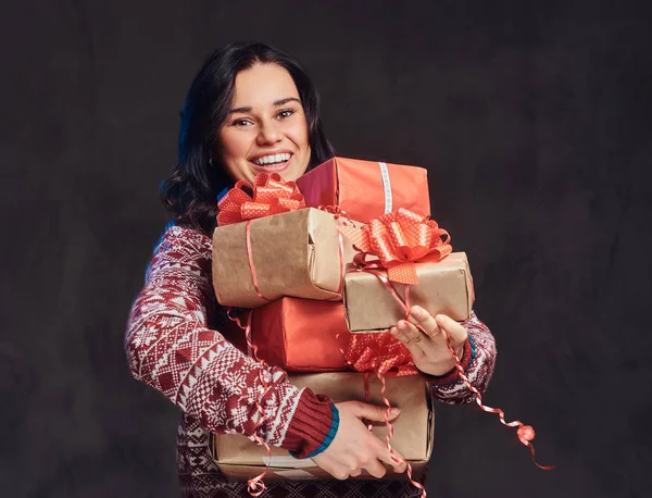 Retrato de uma menina morena feliz vestindo uma camisola quente segurando uma caixas de presentes, isolado em um fundo escuro texturizado . — Fotografia de Stock