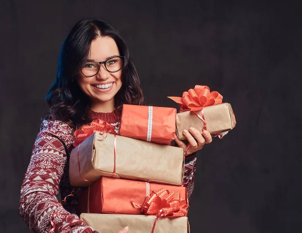 Retrato de uma menina morena feliz vestindo óculos e suéter quente segurando uma caixas de presentes, isolado em um fundo escuro texturizado . — Fotografia de Stock