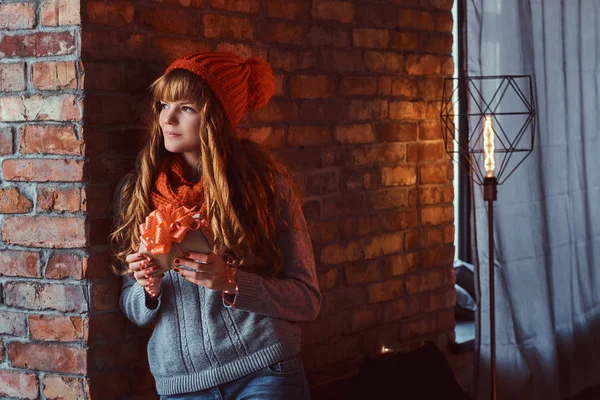 Portret van een eenzame roodharige meisje draagt een warme trui en muts een doos van de gift te houden terwijl leunend op een bakstenen muur. — Stockfoto