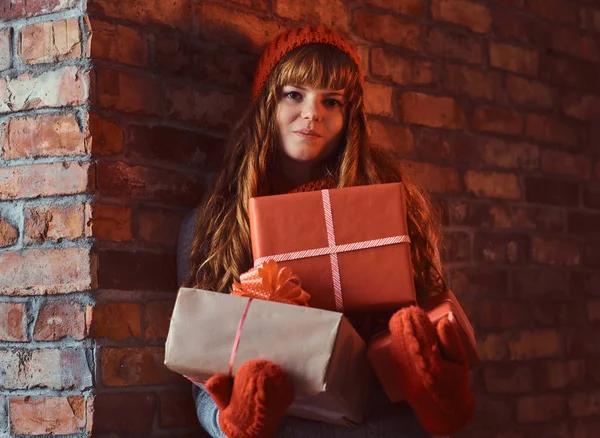 Portrait of a lonely redhead girl wearing a warm sweater and hat holding gift boxes while leaning on a brick wall. — Stock Photo, Image