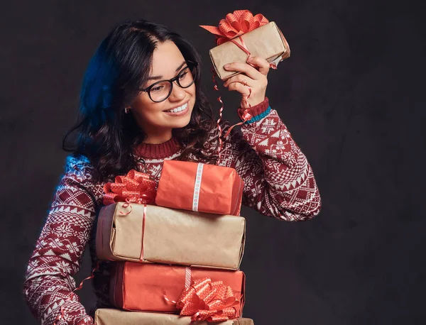 Portrait of a happy brunette girl wearing eyeglasses and warm sweater holding a gifts boxes, isolated on a dark textured background. — Stock Photo, Image