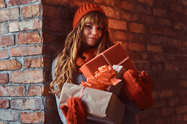 Portrait of a lonely redhead girl wearing a warm sweater and hat holding gift boxes while leaning on a brick wall. — Stock Photo, Image