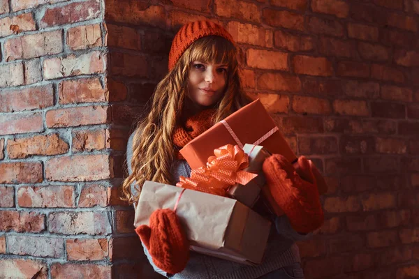 Portrait of a lonely redhead girl wearing a warm sweater and hat holding gift boxes while leaning on a brick wall. — Stock Photo, Image