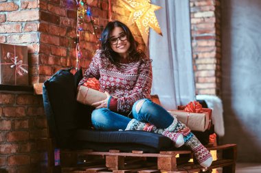 Çatı katı iç ile dekore edilmiş bir odada hediye kutuları ile bir kanepede oturan neşeli güzel kız.