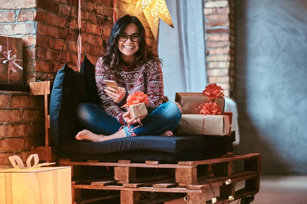 Cheerful beautiful girl holding a phone while sitting on a couch with gift boxes in a decorated room with loft interior. — Stock Photo, Image