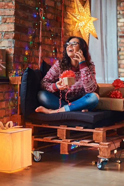 Cheerful beautiful girl talking by phone with friends while sitting on a couch with gift boxes in a decorated room with loft interior. — Stock Photo, Image