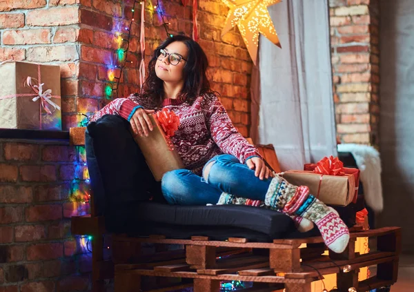 Felice bella ragazza godendo mattina di Natale mentre seduto su un divano con scatole regalo in una stanza decorata con interni soppalco . — Foto Stock
