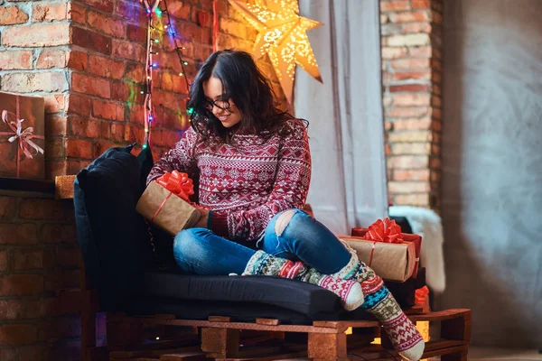 Happy beautiful girl enjoying Christmas morning while sitting on a couch with gift boxes in a decorated room with loft interior. — Stock Photo, Image