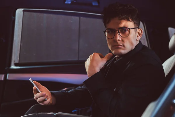 Un joven hombre de negocios guapo sosteniendo un teléfono inteligente mientras está sentado en el asiento trasero del coche de lujo . — Foto de Stock