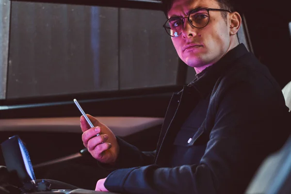 Un joven hombre de negocios guapo sosteniendo un teléfono inteligente mientras está sentado en el asiento trasero del coche de lujo . — Foto de Stock