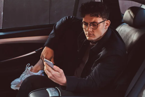 Un joven guapo usando un teléfono inteligente mientras se relaja en el asiento trasero del coche de lujo . — Foto de Stock