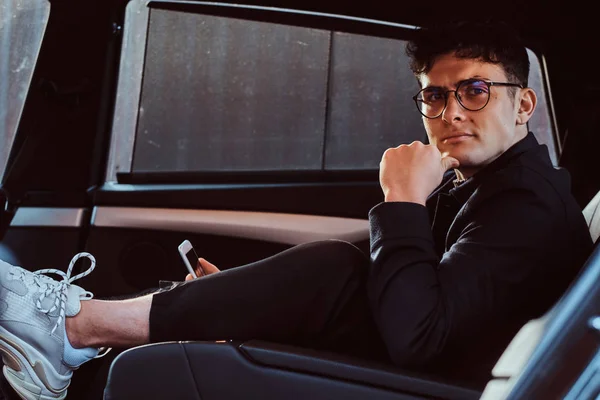 Un joven guapo sosteniendo un teléfono inteligente mientras se relaja en el asiento trasero del coche de lujo . — Foto de Stock