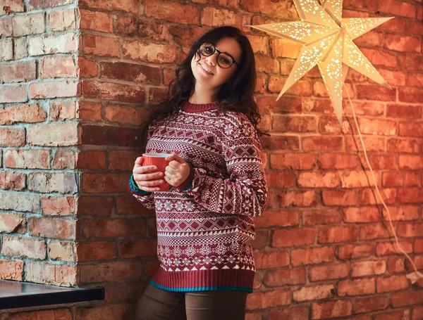 Retrato de uma menina morena sorridente usando óculos e suéter quente segurando uma xícara de café enquanto se inclina em uma parede de tijolo . — Fotografia de Stock