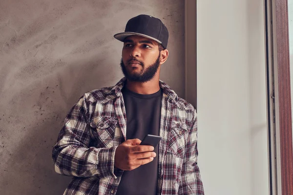 Afroamericano barbudo sosteniendo un teléfono mientras está de pie junto a una pared de textura gris . — Foto de Stock