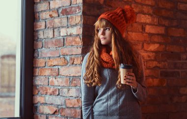 Bir sıcak kazak ve bir fincan kahve bir tuğla duvara yaslanmış ve pencereden dışarı bakarak süre tutarak şapka giyen kızıl saçlı kız.