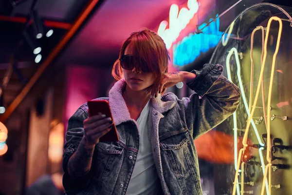 Menina ruiva elegante usando um telefone enquanto se inclina na tabuleta iluminada na noite na rua . — Fotografia de Stock