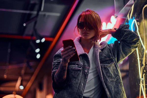 Menina ruiva elegante usando um telefone enquanto se inclina na tabuleta iluminada na noite na rua . — Fotografia de Stock