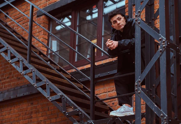 Een modieuze man leunend op een leuning trap buiten het gebouw met de industriële buitenkant. — Stockfoto