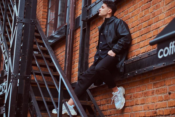 Модний хлопець спирається на стіну, стоячи на сходах за межами будівлі з індустріальним зовнішнім виглядом . — стокове фото