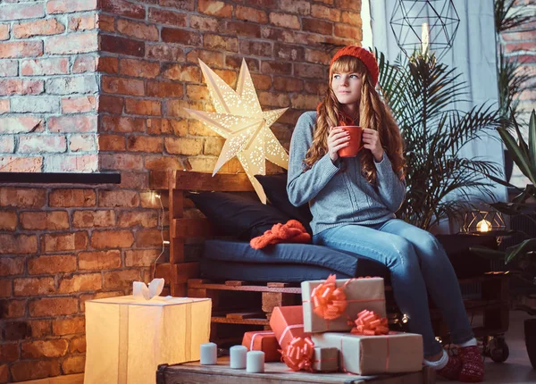红发女郎坐在沙发上, 圣诞节期间在装饰好的客厅里喝咖啡取暖. — 图库照片