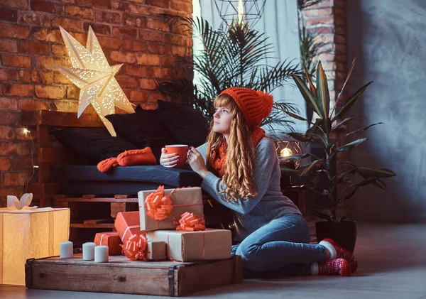 Redhead girl sitting on a floor, warming with a cup of coffee and looking out the window in a decorated living room at Christmas time. — Stock Photo, Image