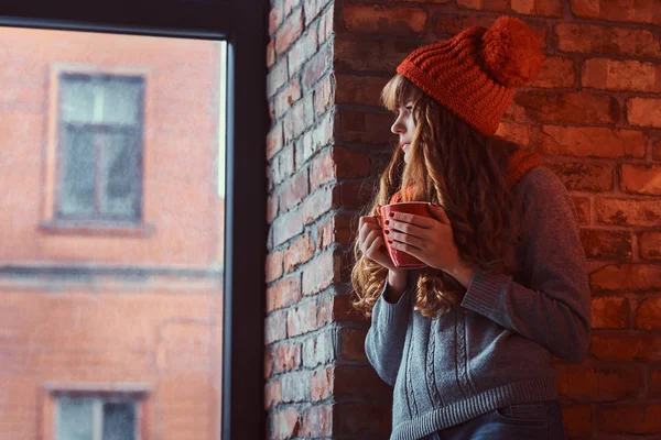 Redhead κορίτσι φορώντας ένα ζεστό πουλόβερ και καπέλο, κρατώντας ένα φλιτζάνι καφέ, ενώ κλίνει σε ένα τοίχο από τούβλα και κοιτάζοντας έξω από το παράθυρο. — Φωτογραφία Αρχείου