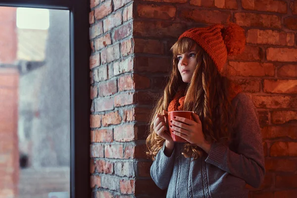 Redhead girl wearing a warm sweater and hat holding a cup of coffee while leaning on a brick wall and looking out the window.