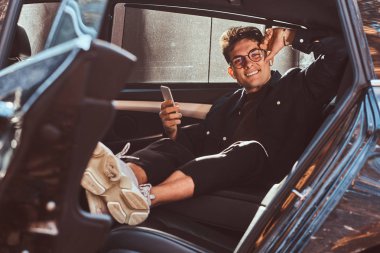 Yakışıklı bir genç adam bir smartphone lüks arabanın arka koltuğunda dinlenirken holding.