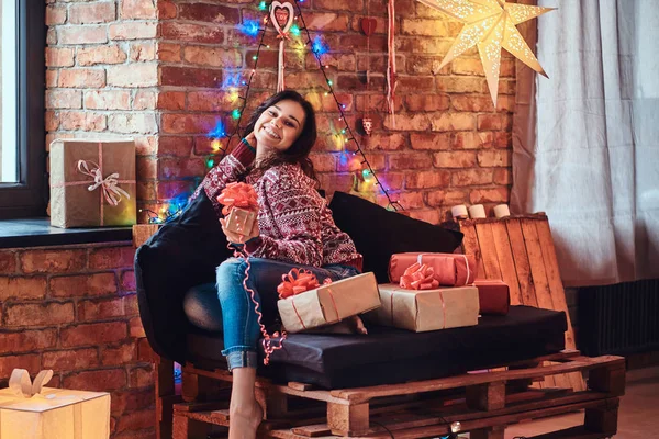 Fröhliche schöne Mädchen sitzt auf einer Couch mit Geschenkboxen in einem dekorierten Raum mit Loft-Interieur. — Stockfoto