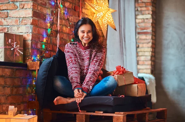 Menina bonita alegre sentado em um sofá com caixas de presente em um quarto decorado com interior loft . — Fotografia de Stock