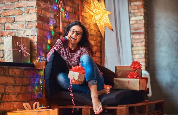 Menina bonita alegre sentado em um sofá com caixas de presente em um quarto decorado com interior loft . — Fotografia de Stock