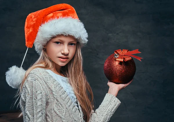 Adolescente usando chapéu Santas segurando uma grande bola de Natal em um fundo escuro texturizado . — Fotografia de Stock