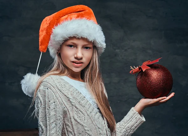 Adolescente usando chapéu Santas segurando uma grande bola de Natal em um fundo escuro texturizado . — Fotografia de Stock