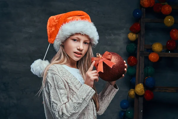 Tiener jongen dragen Santas hoed houden een grote bal van de kerst in een donkere kamer met een versierde houten ladder. — Stockfoto