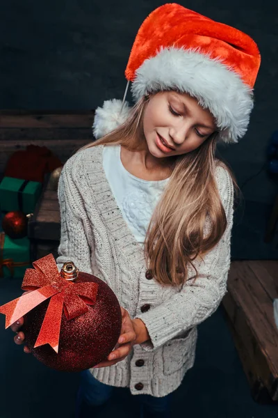 Tiener jongen dragen Santas hoed houden een grote bal van de kerst in een donkere kamer met geschenken. — Stockfoto