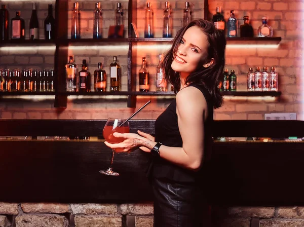 Усміхнена красива дівчина в елегантному одязі тримає коктейль, стоячи поруч з барною стійкою — стокове фото