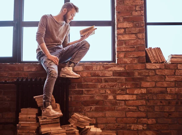 Um jovem estudante bonito com barba elegante e cabelo lendo um livro enquanto sentado em um peitoril da janela — Fotografia de Stock