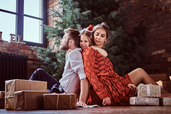 A happy little girl hugging her parents while they sit together on a floor surrounded by gifts next to the Christmas tree at home. — 图库照片