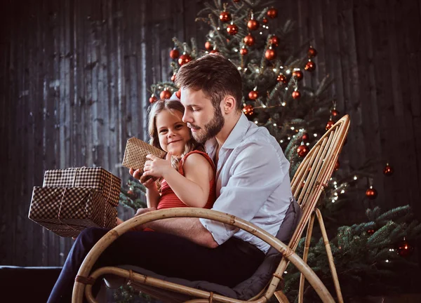 Baba ve kızı birlikte bir sallanan sandalyede otururken hediye kutuları tutarak bir Noel ağacı evde yakın:. — Stok fotoğraf
