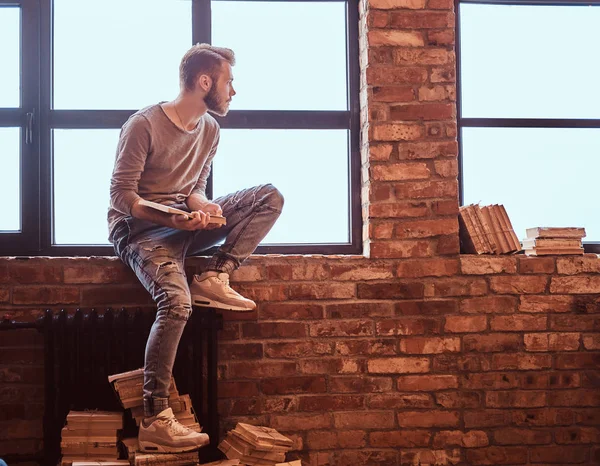 A young handsome student with stylish beard and hair holding a book and looking out the window — Stock Photo, Image