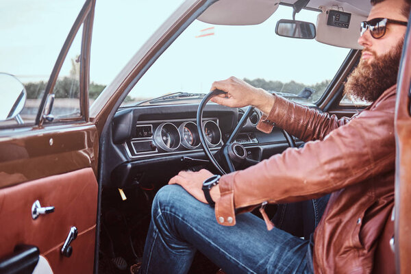 Bearded male in sunglasses dressed in brown leather jacket sits behind the wheel of a tuned retro car with open door.