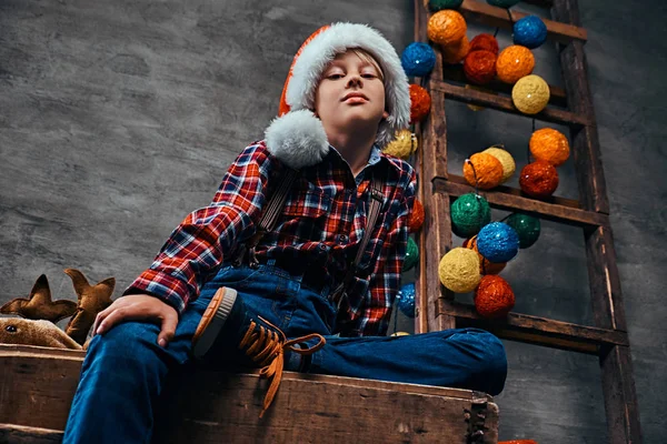 Jeune garçon en chapeau Santas portant une chemise à carreaux avec des bretelles assises sur une palette à côté d'une échelle décorée en bois . — Photo