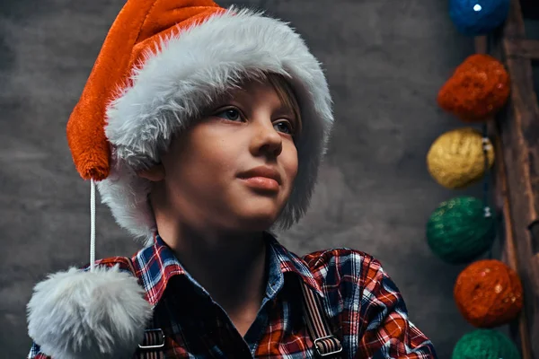 Un ragazzino col cappello di Babbo Natale che indossa una camicia a scacchi con le bretelle. Isolato su uno sfondo scuro strutturato . — Foto Stock