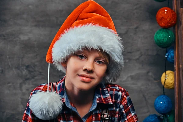 Tiener jongen in Santas hoed dragen een geruite hemd met jarretels. Geïsoleerd op een donkere gestructureerde achtergrond. — Stockfoto