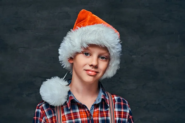 A handsome teen boy in Santas hat wearing a checkered shirt with suspenders. Isolated on a dark textured background. — Stock Photo, Image